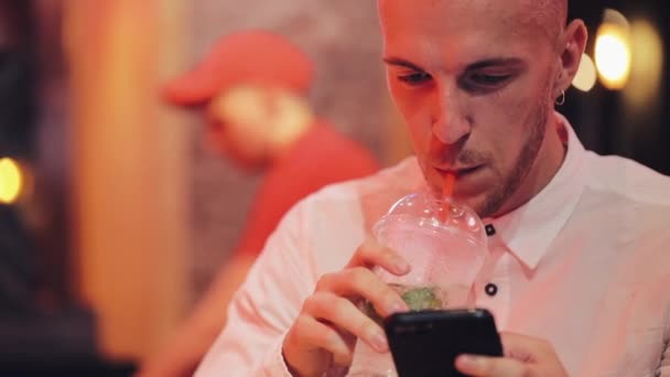 Akşam saatlerinde Smartphone kullanan genç çekici adam. O, bir bar veya restoran neon tabela yakın oturan ve kokteyl içme. İletişim, dinlenme, sohbet, seyahat kavramı. Yakın çekim — Stok video