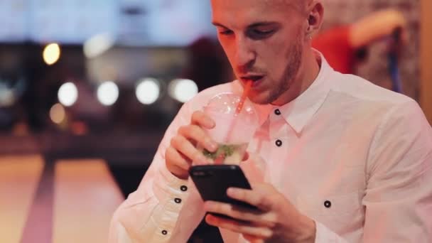 Joven hombre atractivo usando Smartphone en la noche. Se sienta en un bar o restaurante cerca de la señalización de neón y beber cóctel. Comunicación, descanso, chat, concepto de viaje. De cerca. — Vídeo de stock