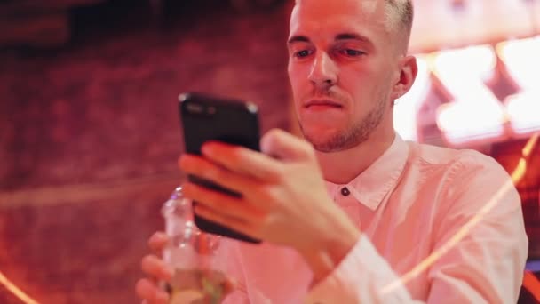Mladý atraktivní muž pomocí Smartphone na večer. Sedí v baru nebo restauraci poblíž neonové poutače a pít koktejl. Komunikace, odpočinku, chatování, cestování koncept. Detailní záběr — Stock video