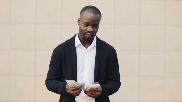 Retrato de um empresário afro-americano de fato a contar dinheiro perto do centro de escritórios. Ele comemorando sua vitória bem sucedida com um monte de dólares e olhando para a câmera — Vídeo de Stock