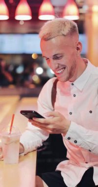 Dikey video. Genç oturuşta bar, içki limonata ve neon bar işareti yanında onun smartphone kullanarak adam. Müjde müjde diğerlerine. Kazançlar, piyango, iyi haber kavramı