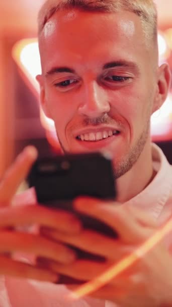 Vertical video. Young Attractive Man with Beautiful Smile is Using Smartphone at Evening. He sitting in a bar or restaurant near neon signage. Close up, 4k. Vertical frame for mobile devices — Stock Video