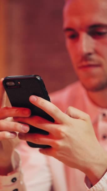 Vídeo vertical. Joven hombre atractivo usando Smartphone en la noche. Está sentado en un bar o restaurante cerca de la señalización de neón. Cierra, 4k. Marco vertical para dispositivos móviles — Vídeos de Stock