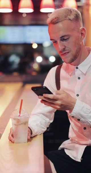 Pionowe wideo. Młody człowiek siedzi w barze, picia lemoniady i za pomocą jego smartphone obok neon znak bar. Turystycznych, ciesząc się nocne życie podczas komunikowania się z przyjaciółmi na swoim smartfonie — Wideo stockowe