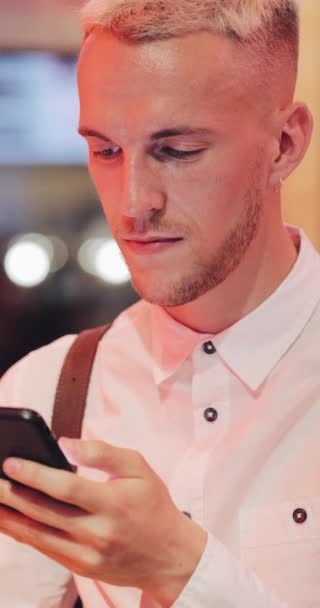 Vídeo vertical. Joven sentado en el bar, bebiendo limonada y usando su teléfono inteligente junto al letrero del bar de neón. Turista disfrutando de la vida nocturna mientras se comunica con amigos en su teléfono inteligente — Vídeo de stock