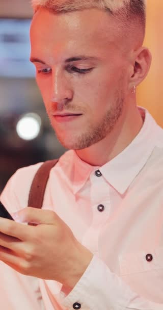 Vidéo verticale. Jeune homme heureux en utilisant smartphone dans un café confortable à l'heure du soir. Communication, affaires, voyage, sortir ensemble concept — Video