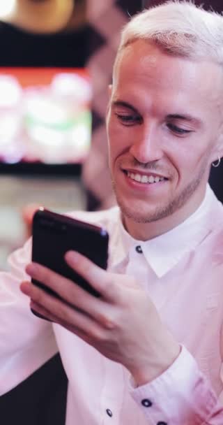 Вертикальное видео. Молодой счастливый человек использует смартфон в уютном кафе в вечернее время. Концепция знакомств, бизнес, путешествия — стоковое видео