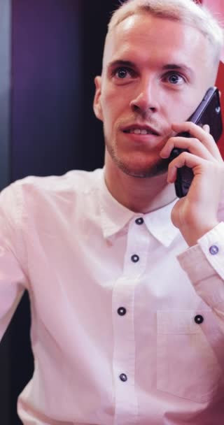 Vertical video. Young handsome man talking on the smartphone in the night cafe. He sitting near neon signage. Communication and business concept — Stock Video
