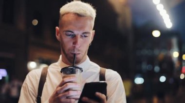 Gülümseyen adam usingn smartphone gece şehirde. Geceleri Smartphone kullanarak ve buzlu kahve içme yakışıklı bir genç adam. Hareket eden iletişim, iş, seyahat kavramı
