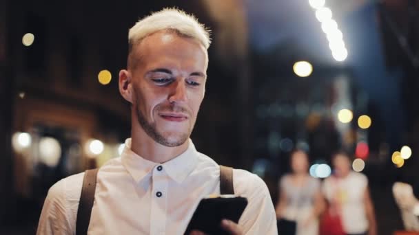 Uomo sorridente che utilizza uno smartphone nella città di notte. Bel giovanotto che usa lo smartphone di notte. Comunicazione di mobilità, business, concetto di viaggio — Video Stock