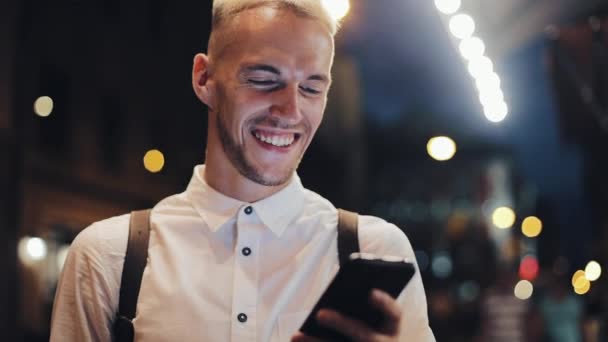 Uśmiechnięty mężczyzna usingn smartfona w nocy miasto. Przystojny młody człowiek za pomocą smartfona w nocy i picia kawy lodu. Mobilności komunikacja, biznes, koncepcja podróży — Wideo stockowe