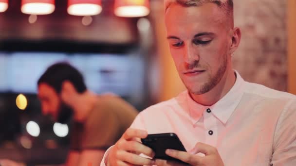 Mladý pohledný muž vytočit číslo a mluví o smartphone v noční kavárně. On sedí poblíž neonové poutače. Komunikační a obchodní koncept — Stock video