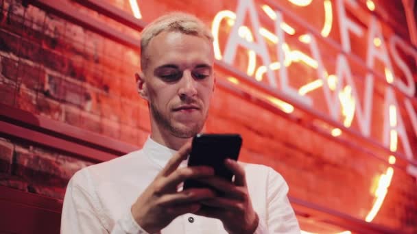 Giovane uomo attraente utilizzando Smartphone alla sera. Si trovava in un bar o in un ristorante vicino alla segnaletica al neon. Comunicazione, riposo, chat, concetto di viaggio. Da vicino. — Video Stock