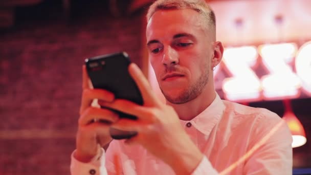 Joven hombre atractivo usando Smartphone en la noche. Está sentado en un bar o restaurante cerca de la señalización de neón. Comunicación, descanso, chat, concepto de viaje. De cerca. — Vídeos de Stock