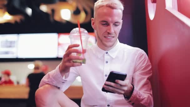 Jovem Homem Atraente Usando Smartphone à noite. Ele sentado em um bar ou restaurante perto de sinalização de néon e beber coquetel. Comunicação, descanso, conversação, conceito de viagem — Vídeo de Stock