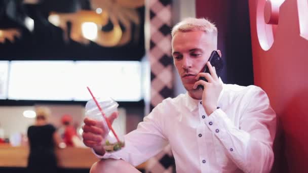 Молодой красивый мужчина разговаривает по смартфону в ночном кафе. Он сидит рядом с неоновой вывеской и пьет коктейль. Коммуникация и бизнес-концепция — стоковое видео