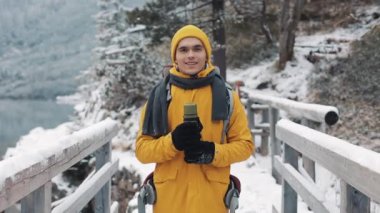 Kış giysileri bir termos elinde olan genç bir çekici adam portresi. Sarı kış giysileri giyen yürüyüş adam kamera ve gülümsüyor görünüyor. Bir kar köprü üzerinde durduğunu