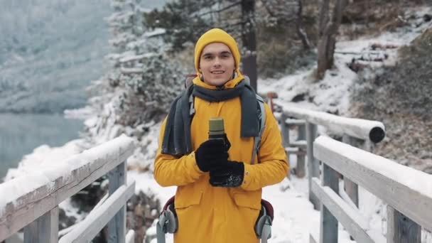 Porträtt av en ung attraktiv man i vinterkläder med en termos i handen. Vandring man klädd i gul vinterkläder ser på kameran och leenden. Han står på en snö-bro — Stockvideo