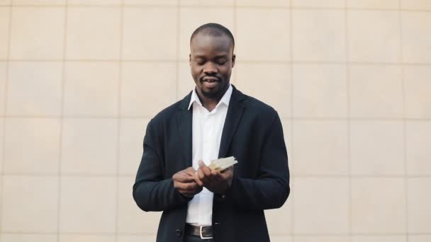 Homem de negócios afro-americano feliz com uma grande quantidade de dinheiro nas mãos. Ele fica na rua perto do centro de escritório ou banco e mostra dinheiro para a câmera — Vídeo de Stock