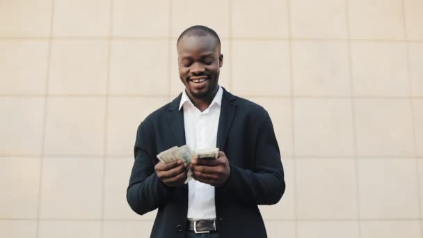 Портрет африканского американского бизнесмена в костюме, который считает деньги, стоящие рядом с офисным центром. Он празднует свою победу с кучей долларов и смотрит в камеру — стоковое видео