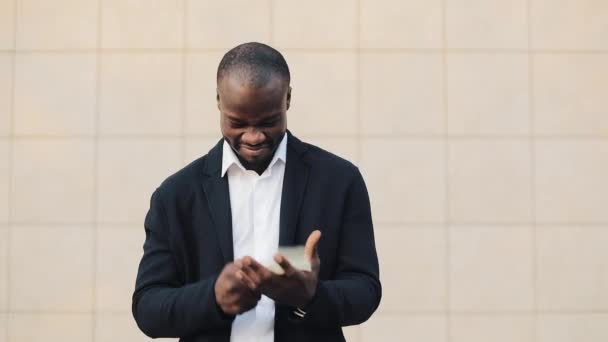 Porträt eines afrikanisch-amerikanischen Geschäftsmannes im Anzug, der Geld zählt, das in der Nähe eines Bürozentrums steht. er feiert seinen erfolgreichen Sieg mit vielen Dollars und blickt in die Kamera — Stockvideo