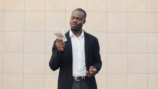 Retrato de um empresário afro-americano de fato a contar dinheiro perto do centro de escritórios. Ele comemorando sua vitória bem sucedida com um monte de dólares e olhando para a câmera — Vídeo de Stock