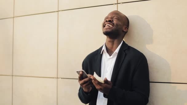 Gelukkig Afro-Amerikaanse zakenman juichen vieren kijken naar mobiele telefoon en een grote hoeveelheid geld in zijn handen houden. Hij staat in de straat in de buurt van het office-center of bank — Stockvideo