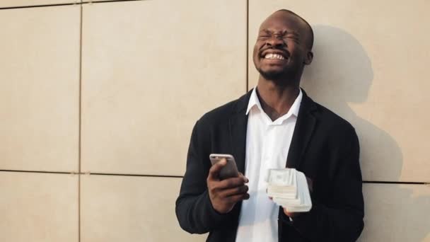 Un om de afaceri african fericit, care sărbătorește uitându-se la telefonul mobil și ținând în mână o sumă mare de bani. Stă pe stradă lângă centrul de birouri sau bancă. — Videoclip de stoc