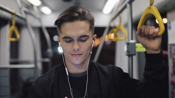 Portrait of young headsome men in headphones listening to music and browsing on mobile phone in public transport. City lights background. Slow motion — Stock Video