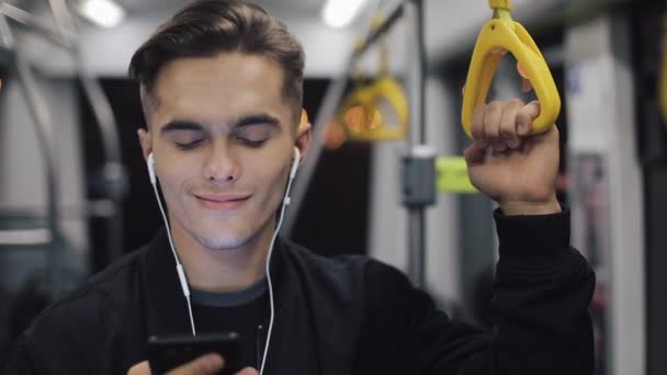 Retrato de hombres jóvenes en auriculares escuchando música y navegando en el teléfono móvil en el transporte público. Fondo de luces de ciudad. Movimiento lento — Vídeo de stock