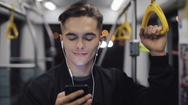 Портрет молодого человека в наушниках, слушающего музыку и просматривающего мобильный телефон в общественном транспорте. Городские огни фона. Медленное движение — стоковое видео