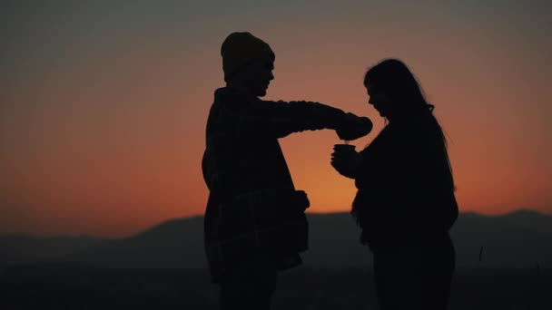 Silueta de pareja joven enamorada de beber té de una taza de termos en el hermoso fondo de la puesta de sol de las montañas. Turismo, cita romántica, descanso, vacaciones, naturaleza, concepto de caminata — Vídeos de Stock