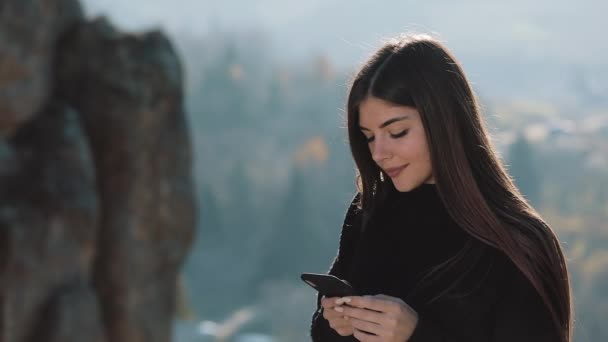 若い幸せな女は 美しい山の前にある岩の上に彼の電話の立場で誰かとチャットします ハイキングのコンセプト — ストック動画