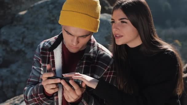 Viaja a las montañas. Hermosa pareja enamorada se sienta en la roca y el uso de mapa en el teléfono inteligente. Hombre muestra novia algo en su teléfono . — Vídeo de stock