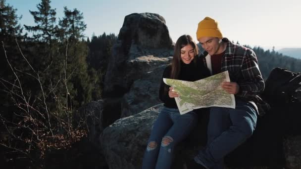 Поезжай в горы. Красивая влюбленная пара сидит на скале и изучает карту. Они разговаривают друг с другом, смеются и веселятся — стоковое видео