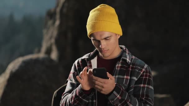 Junger glücklicher Mann plaudert mit jemandem auf seinem Handy, der auf der Spitze des Felsens vor dem schönen Berg steht. Kommunikation, Tourismus, Wanderkonzept — Stockvideo