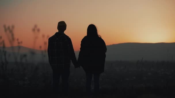 Silueta de pareja joven enamorada disfrutando de una puesta de sol sobre las montañas. Vacaciones, viajes, romance, concepto de propuesta de matrimonio — Vídeos de Stock