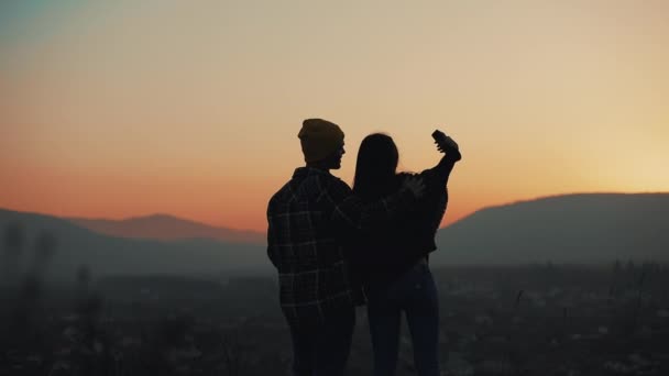 Silueta de pareja joven enamorada disfrutando de una puesta de sol sobre las montañas. Toman fotos en un smartphone. Vacaciones, concepto de viaje — Vídeo de stock