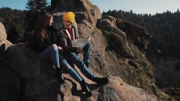 Поезжай в горы. Красивая влюбленная пара сидит на скале и изучает карту. Они разговаривают друг с другом, смеются и веселятся — стоковое видео