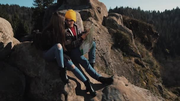 Travel to the mountains. Beautiful couple in love sits on the rock and learns a map. They talk with each other, laughing and having fun — Stock Video