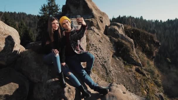 Jovem casal apaixonado senta-se na rocha e tirar um retrato selfie. Homem e mulher visitando atração turística famosa — Vídeo de Stock