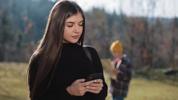 Wanita muda cantik berdiri di dekat pegunungan menggunakan smartphone-nya, pesan mengetik. Turis dengan peta di latar belakang — Stok Video