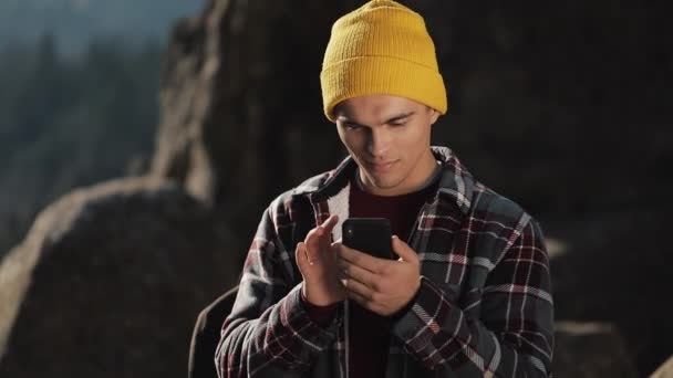 Joven hombre feliz charla con alguien en su teléfono de pie en la cima de la roca antes de la hermosa montaña. Comunicación, turismo, concepto de caminata — Vídeos de Stock