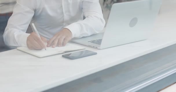 Der junge Geschäftsmann notiert Geschäftsinformationen in seinen Notizblock, während er im gläsernen Café auf seinen Laptop blickt. Zeitlupe, Mittagszeit — Stockvideo