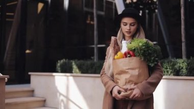 Genç güzel kadın bir paket gülümseyen ve etrafa ürünlerin holding sokakta yürürken şık bir palto giyiyor. Alışveriş, sağlıklı beslenme