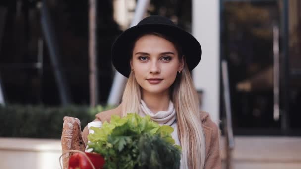 Mujer hermosa joven con un abrigo elegante de pie en la calle sosteniendo un paquete de productos sonriendo y mirando a la cámara. Compras, alimentación saludable — Vídeo de stock