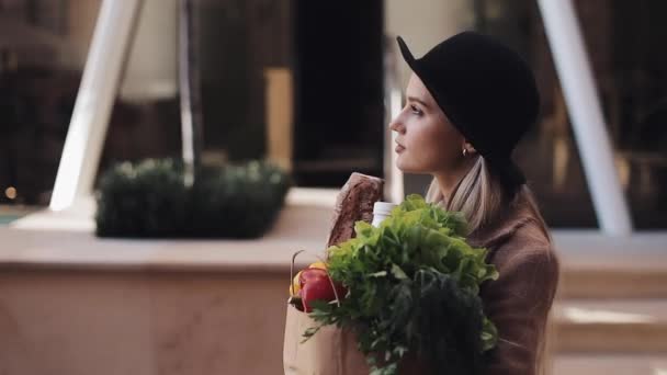 Mladá krásná žena nosí stylový kabát chůzi na ulici, drží balíček produktů, usmíval se a rozhlížel se kolem. Nakupování, zdravé stravování — Stock video