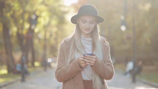スタイリッシュなコートと秋の市の公園で散歩中にスマート フォンを使用して黒の帽子を身に着けている若い美しい女性 — ストック動画