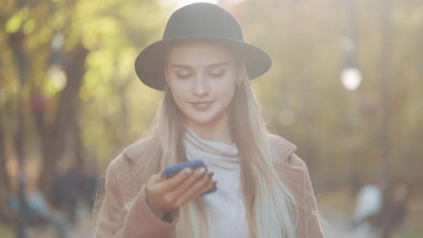 Mujer hermosa joven con abrigo elegante y sombrero negro caminando en el parque de otoño utilizando el mensaje de voz en el teléfono inteligente — Vídeos de Stock