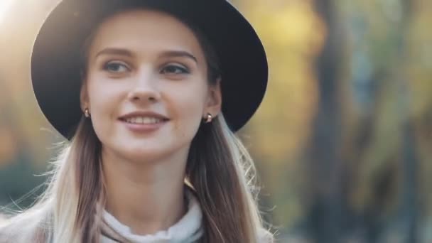 Menina incrível em um olhar romântico, com chapéu preto andando pelo beco do parque, olhando ao redor e dá um sorriso incrível. Configurações do parque, árvores, folhas caídas no fundo — Vídeo de Stock